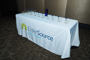A table is placed at the front of a room. It's covered in a white tablecloth that says "ElderSource." Over a dozen awards are placed on the table.