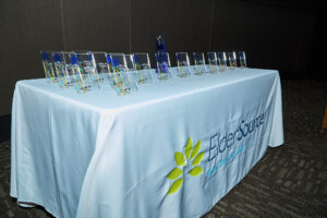 A table is draped in a white tablecloth with the ElderSource logo. Over a dozen awards are arranged on the table.