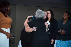 Two women hug while three others stand nearby holding awards.